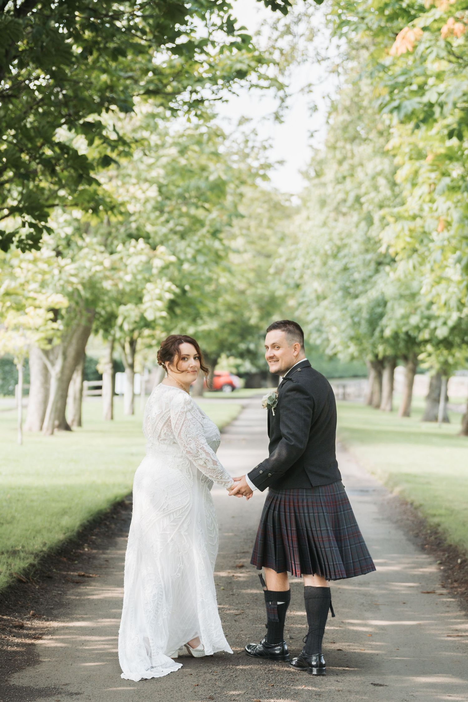 A couple in wedding attire on a tree-lined path, the groom wearing a traditional Scottish kilt and the bride in a lace gown.