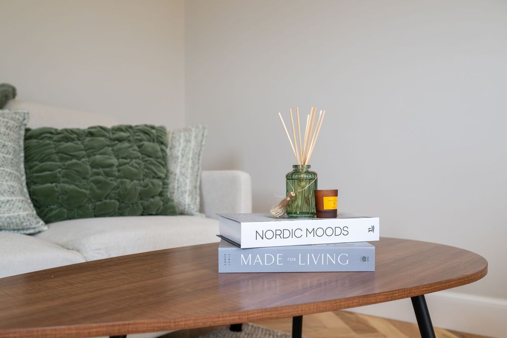 A modern round wooden dining table is styled with a reed diffuser and books in a minimalist interior setting.