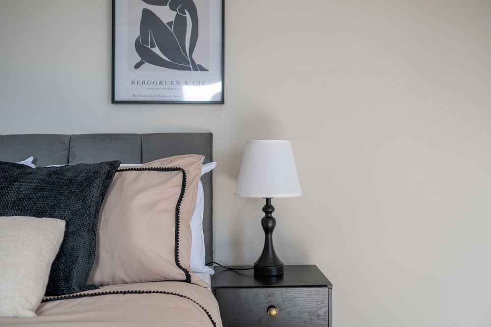 A modern bedroom interior with a black and beige color scheme featuring a bedside lamp and framed artwork on the wall.