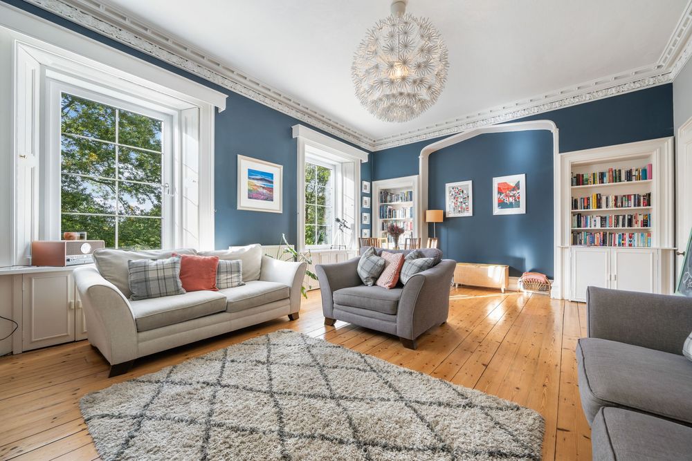 Panoramic view of a modern living room with blue walls, grey sofas, and hardwood flooring under a crystal chandelier.