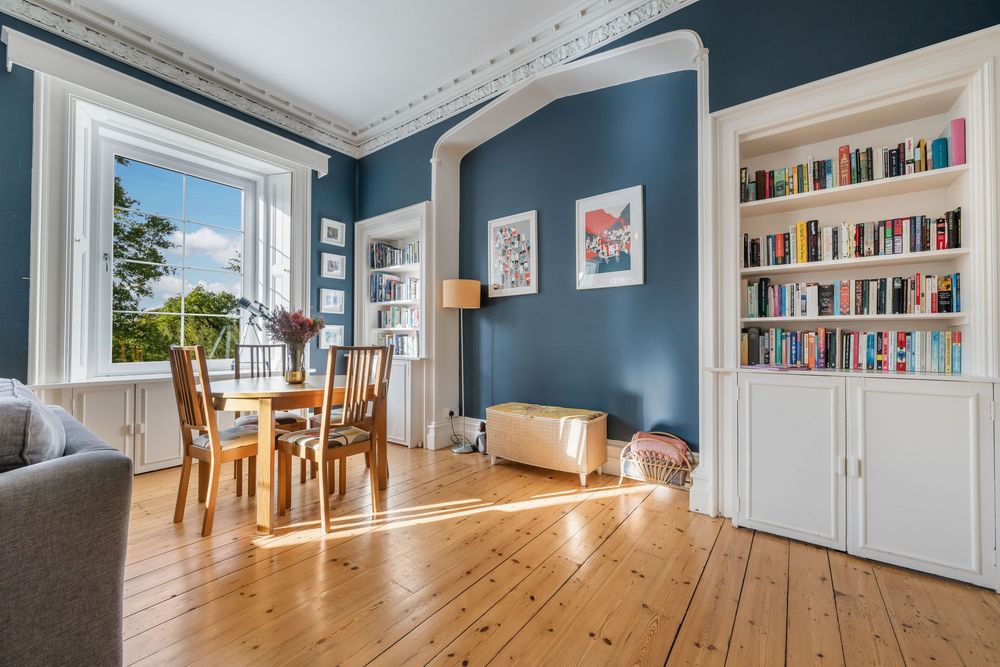 Bright living room with deep blue walls, wooden floors, built-in bookshelves, and dining area near large window.