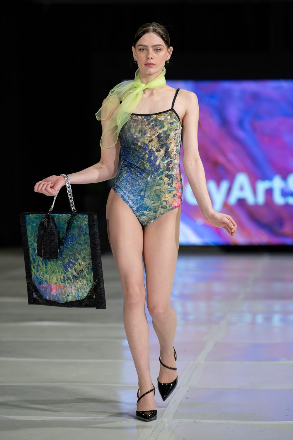 Fashion model walks runway in iridescent swimsuit with matching bag and neon scarf at fashion show.