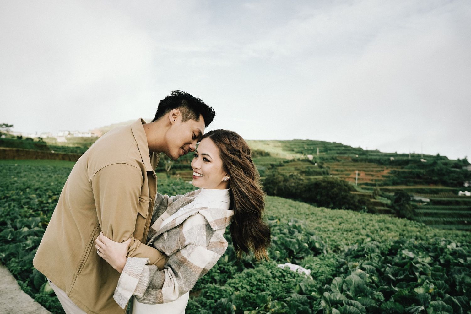 Romantic couple embrace in casual attire against scenic green countryside landscape.