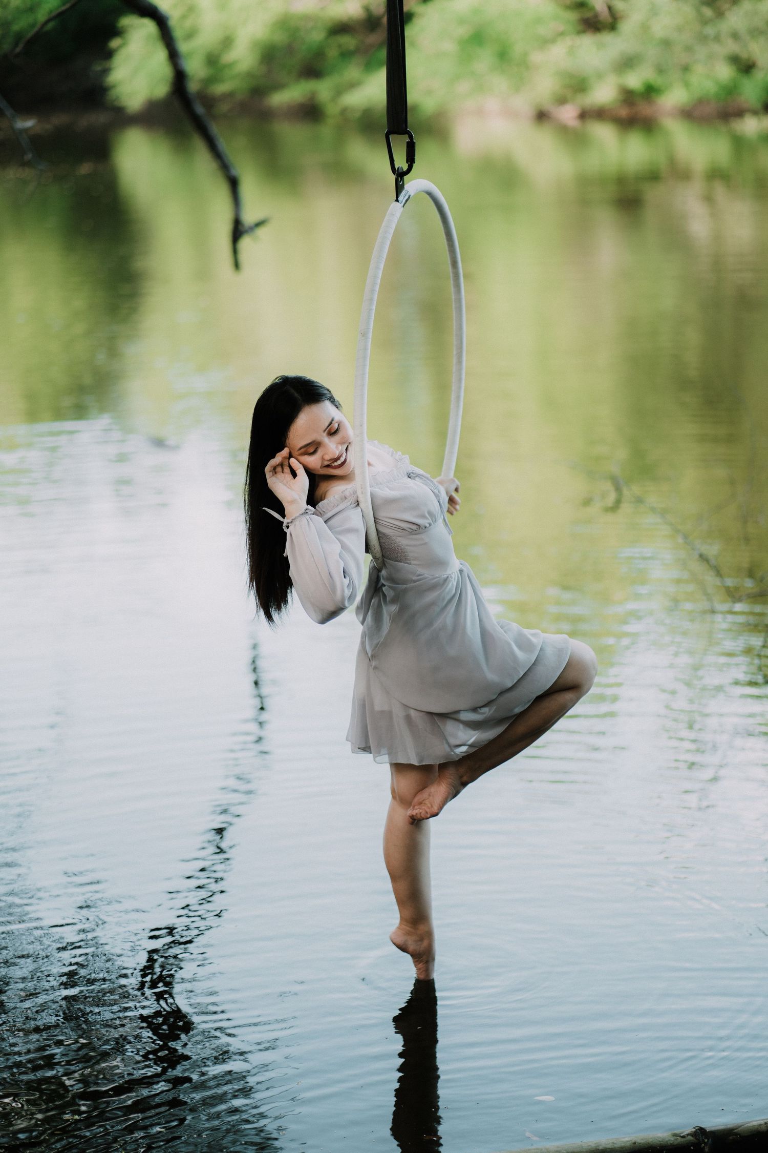 A person in a gray dress poses gracefully on a suspended hoop over water in a natural setting.