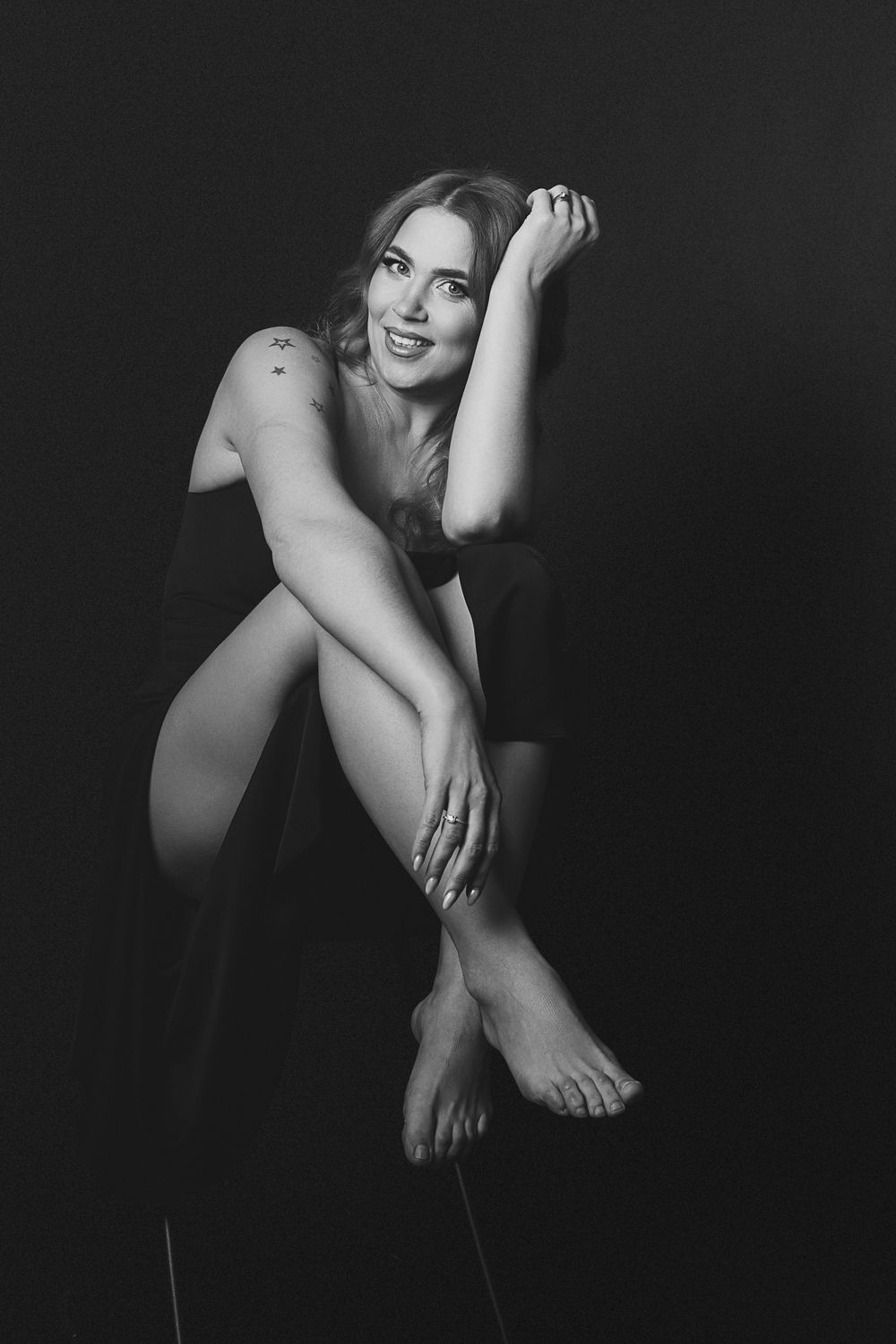 Black and white portrait of a smiling blonde woman sitting barefoot in a dark studio setting.