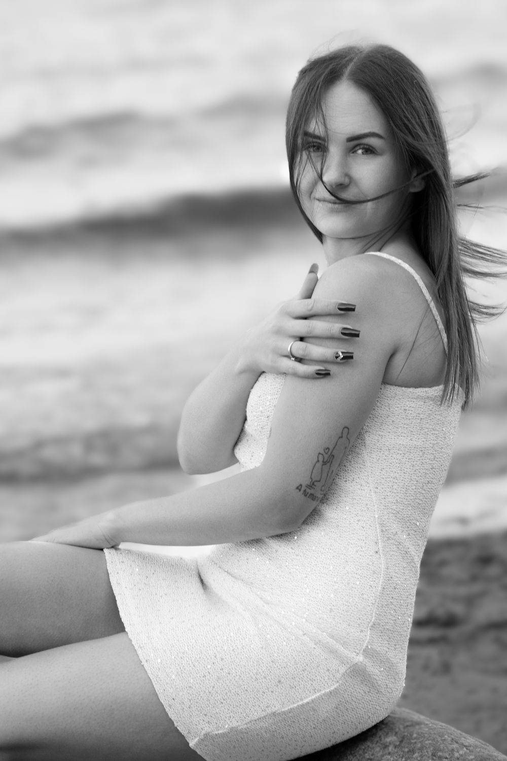 Black and white photo of a young woman in a white dress sitting by the ocean, hair blowing in the wind.