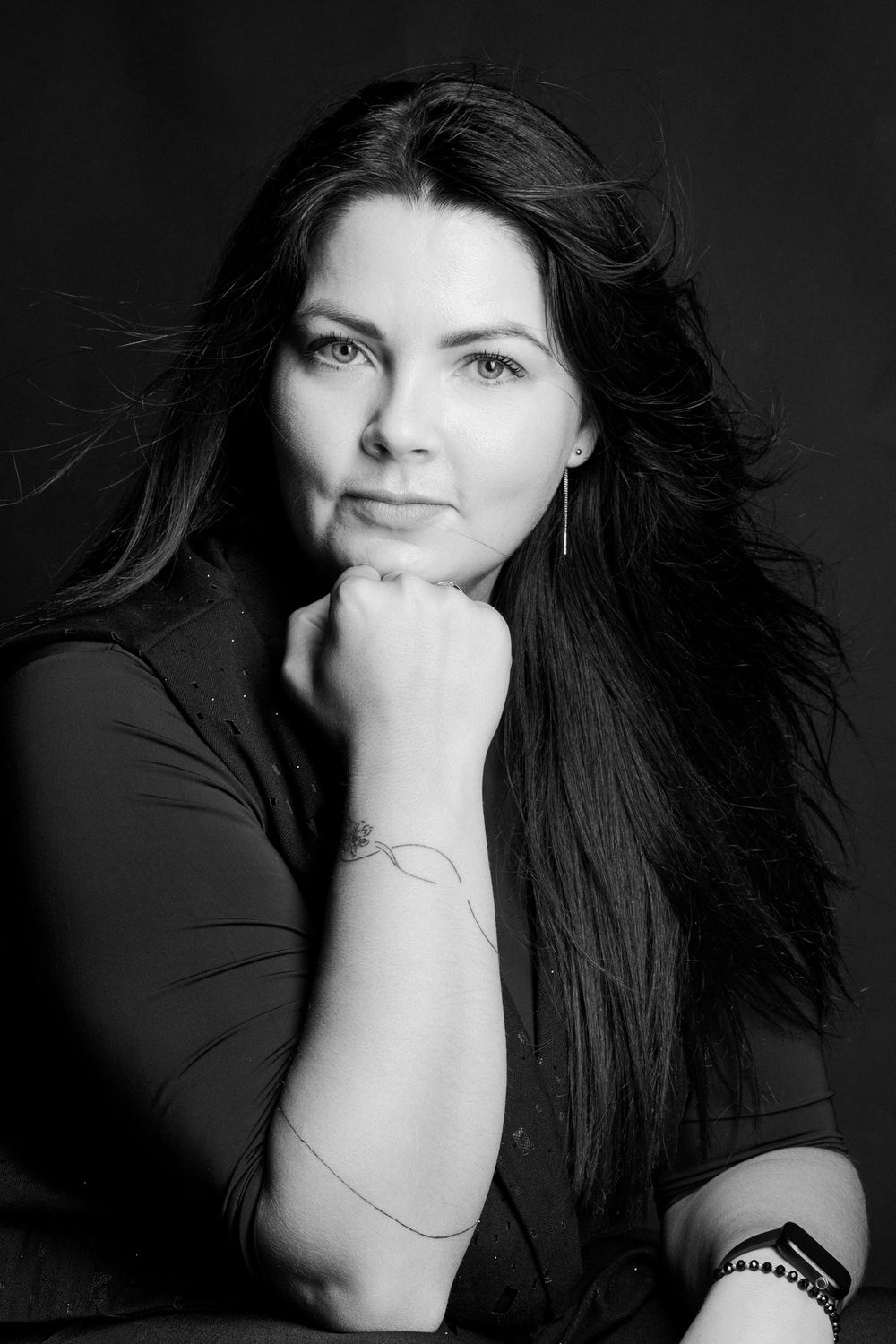 Black and white portrait of a woman with long dark hair resting her chin on her hand against a dark background.