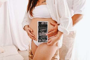 Pregnant woman in white bikini holding ultrasound scan photos while partner embraces her belly in bright room.