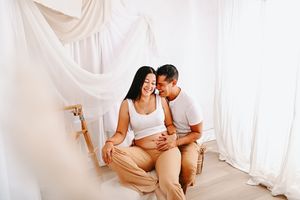 Happy pregnant couple sitting together in a bright white room, man whispering to smiling woman touching her baby bump.