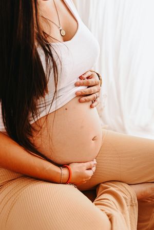 Pregnant woman in white tank top and beige pants cradling her bare baby bump while sitting cross-legged.