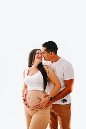 Happy pregnant couple in white tops and tan pants, smiling and embracing while cradling baby bump against white background.