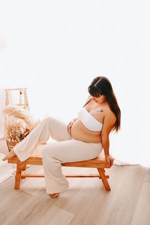 Pregnant woman in white crop top and pants sitting on wooden bench, cradling baby bump in bright minimalist studio.