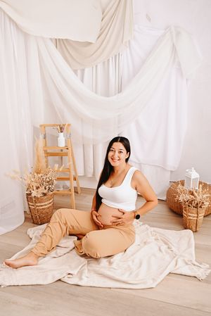 Smiling pregnant woman in white top and beige pants sitting on floor holding baby bump in bright boho studio setting.