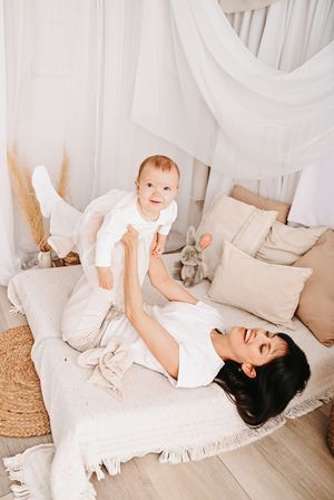 A joyful mother lying on a bed lifts her smiling baby in the air in a cozy, neutral-toned bedroom.