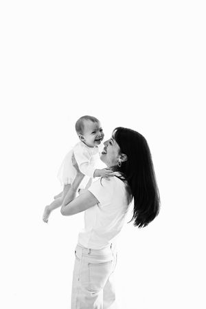 Black and white photo of a joyful mother lifting her laughing baby in the air against a white background.