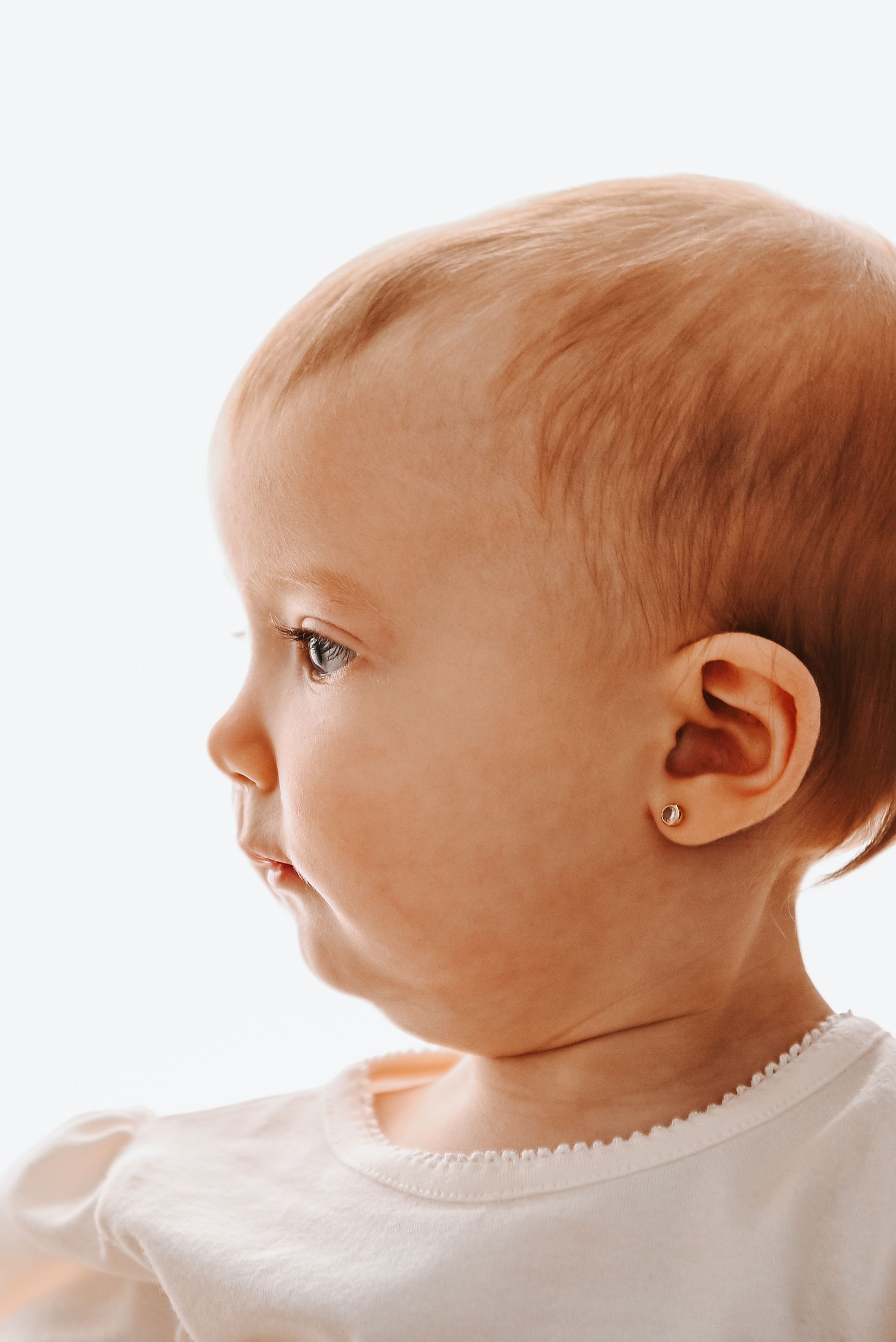 Close-up profile portrait of a baby girl with a small earring, wearing a white onesie against a bright background.
