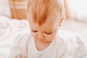 A close-up of an adorable baby with long lashes, wearing a white outfit and pearl necklace, looking down softly.