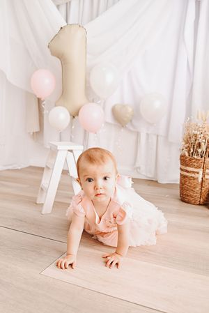 Baby girl in pink dress crawls on floor at first birthday party with number 1 balloon and pastel decorations.
