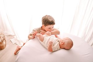 Older toddler boy leaning over and bonding with newborn baby sibling lying on white bed in bright room.