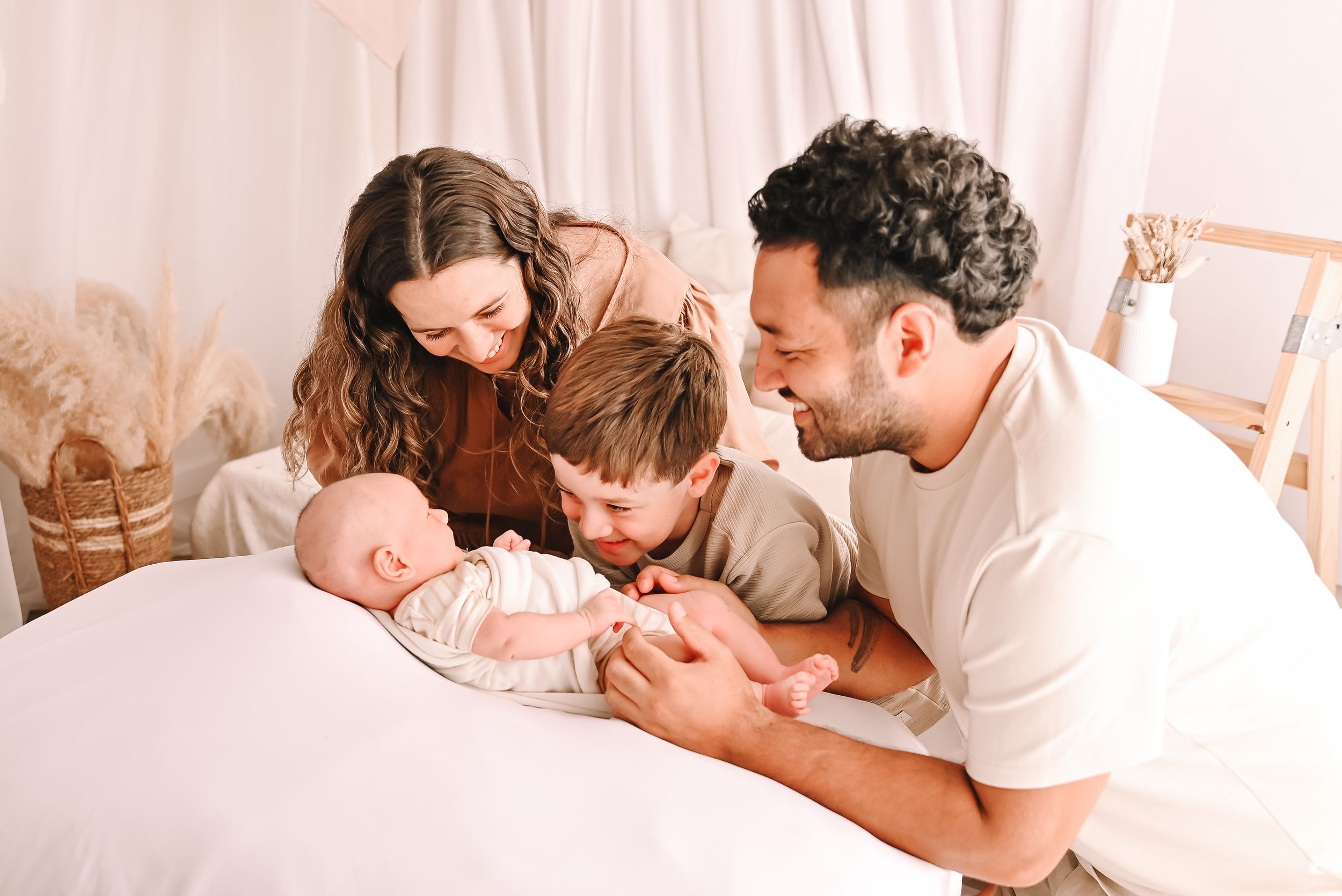 Family of four with newborn baby sharing a tender moment on a white bed in a soft, neutral-toned studio setting.