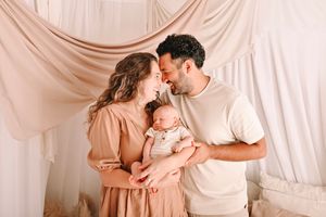 A couple in a beige studio shares a tender moment while holding their newborn baby, surrounded by soft draped fabric.