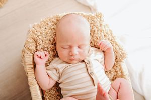 Newborn baby sleeping peacefully in a wicker basket on a soft tan blanket, wearing a striped cream onesie.