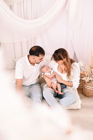 Young parents in white outfits sit together on the floor, lovingly cradling their newborn baby in a soft, bright nursery.