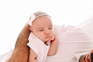Sleeping newborn baby girl in pink outfit and white bow headband cradled in father's arms.