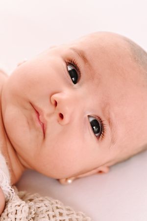 Close-up portrait of a newborn baby with big dark eyes and long lashes lying on a soft white background.