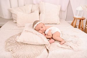 Newborn baby girl sleeping peacefully on a cream pillow with white lace headband in a soft neutral nursery.
