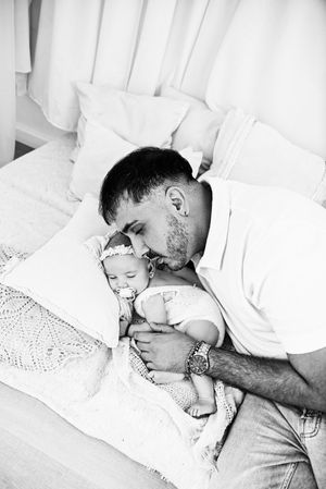 Black and white photo of a father kissing his newborn baby while lying on a bed with white pillows.