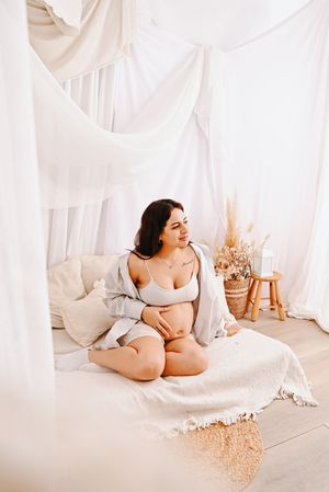 Pregnant woman in white lingerie sitting on a canopy bed, cradling her baby bump in a bright, airy boho bedroom.