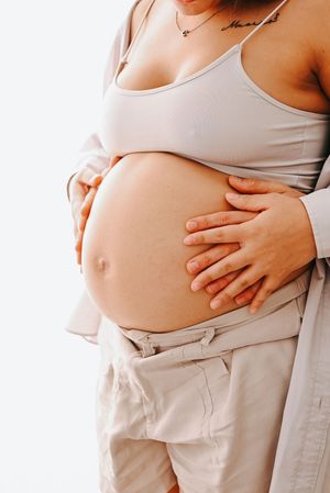 Pregnant woman in beige crop top cradling her bare baby bump with both hands against a white background.