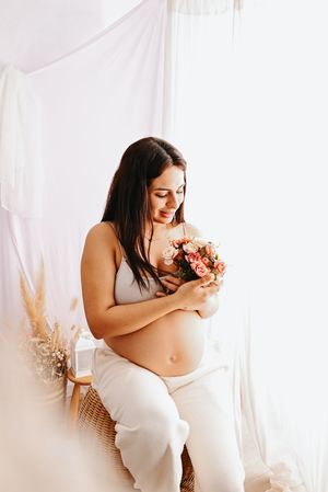Smiling pregnant woman in white crop top holding pink roses bouquet while sitting in wicker chair against soft white backdrop.