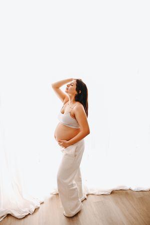 Pregnant woman in gray crop top and white pants posing gracefully near sheer curtains in bright natural light.