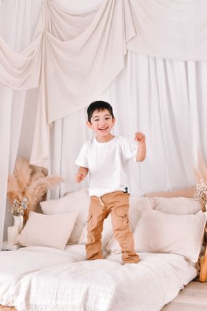 Young child jumping on white mattress against draped fabric backdrop.