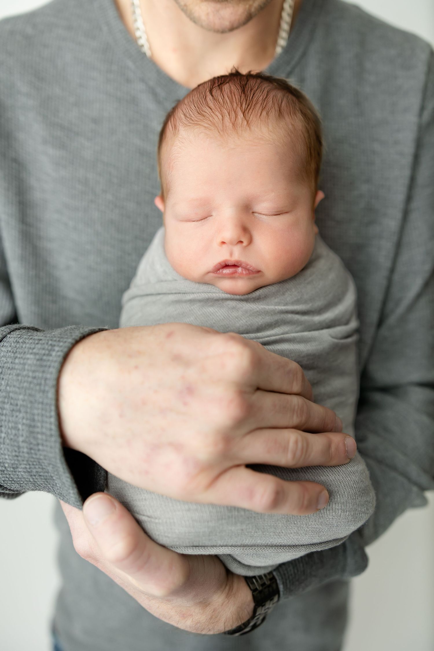 Sleeping newborn wrapped in soft grey fabric being held against person wearing matching grey sweater.