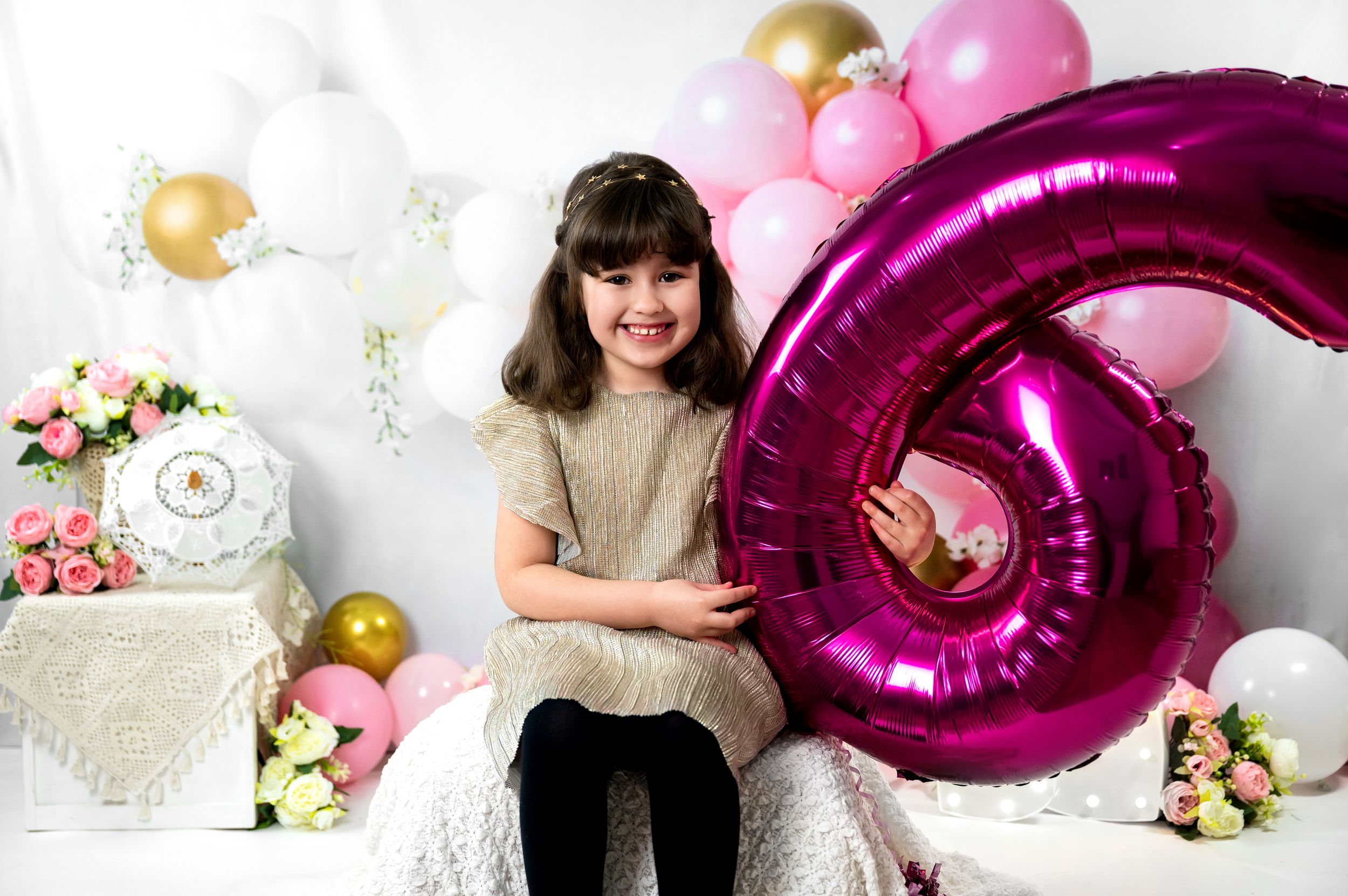 A festive party setup with a large pink number 6 balloon, gold and pink decorations, and floral arrangements against a white backdrop.