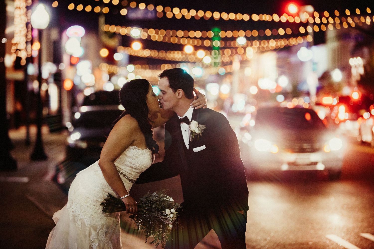 A romantic evening couple shares a moment on city streets with glowing string lights and bokeh in the background.