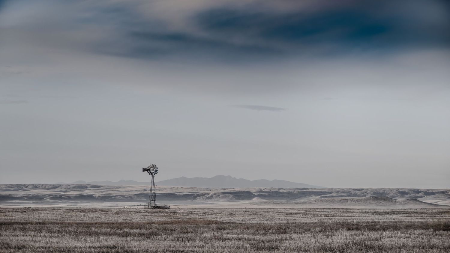 Solitary windmill stands in a vast, empty prairie landscape under moody gray skies.