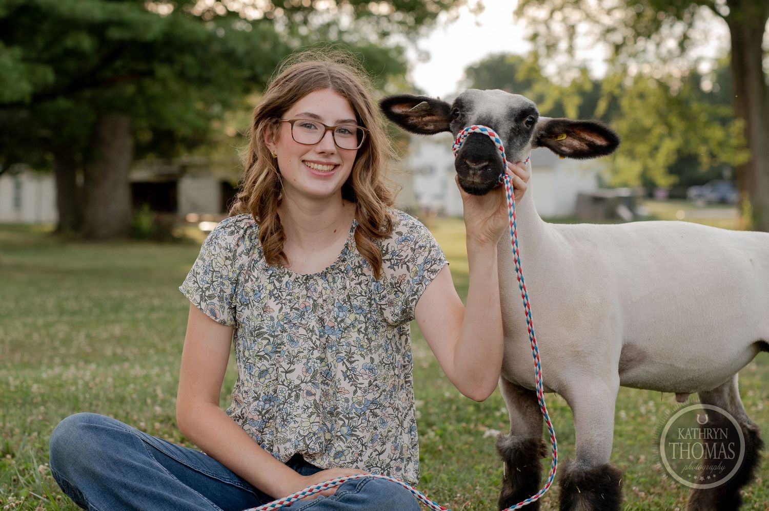A cheerful portrait of a lamb and its handler sitting together in a grassy field with pine trees.