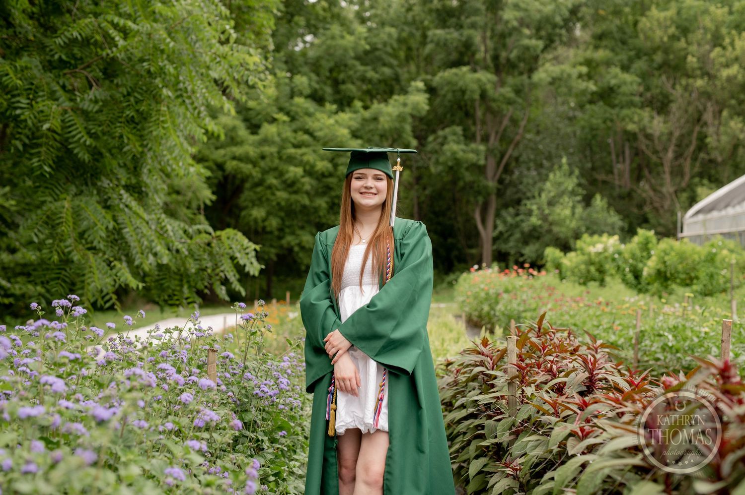 Graduate in emerald green cap and gown stands in garden with purple wildflowers blooming.