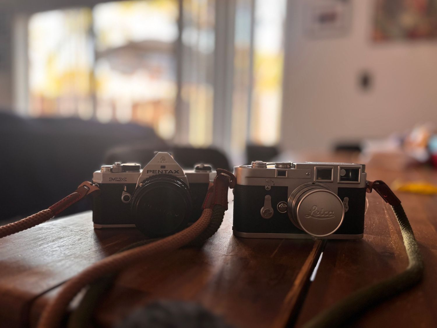 Two vintage cameras with leather straps sit side by side on a brown wooden surface in soft focus lighting.