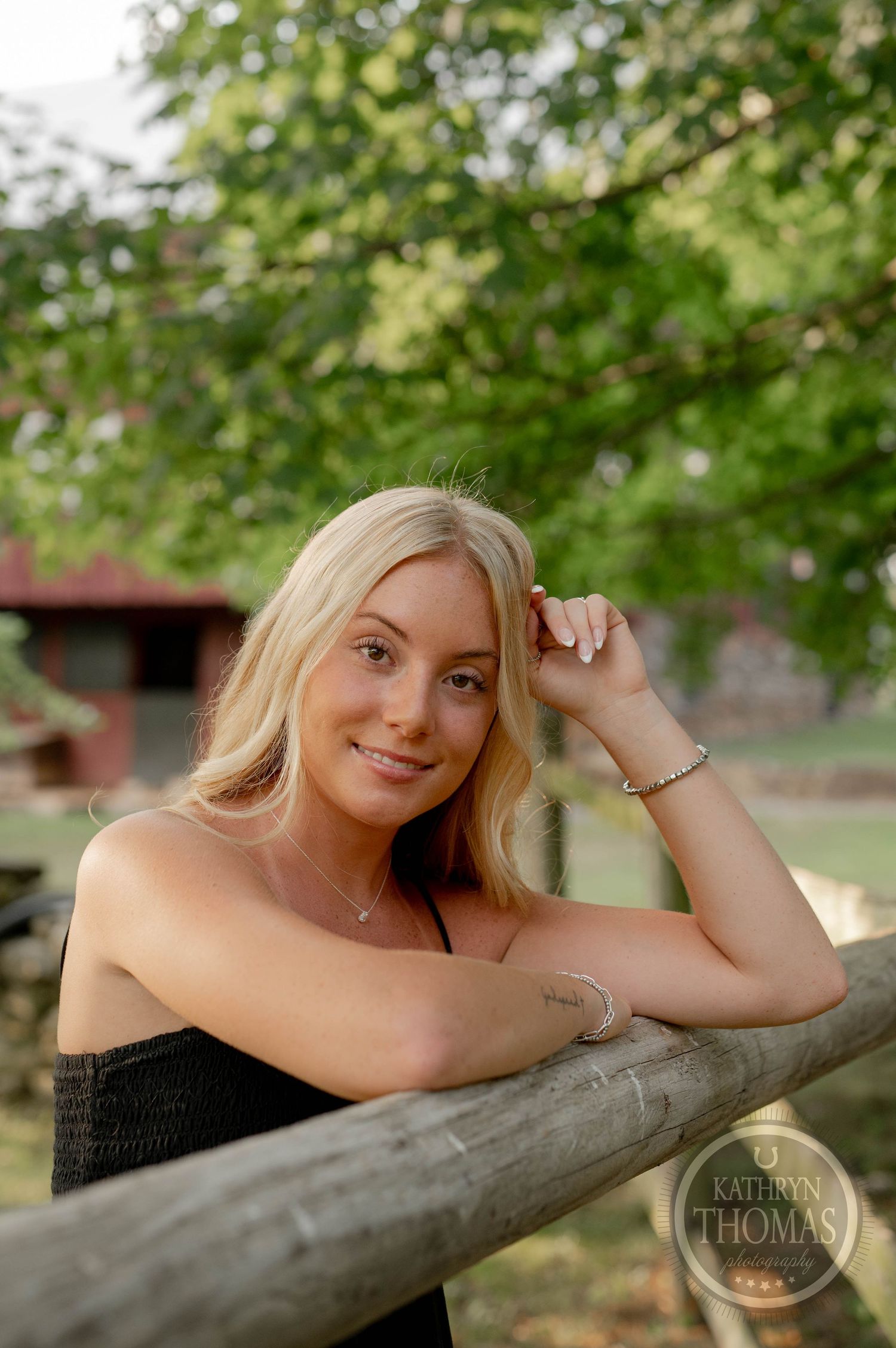 A woman in a black top leans on a rustic wooden fence with trees and greenery behind her.