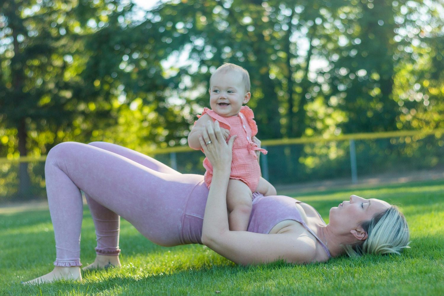 Woman exercising outdoors on grass doing bridge pose while playing with infant in pink outfit on a sunny day.