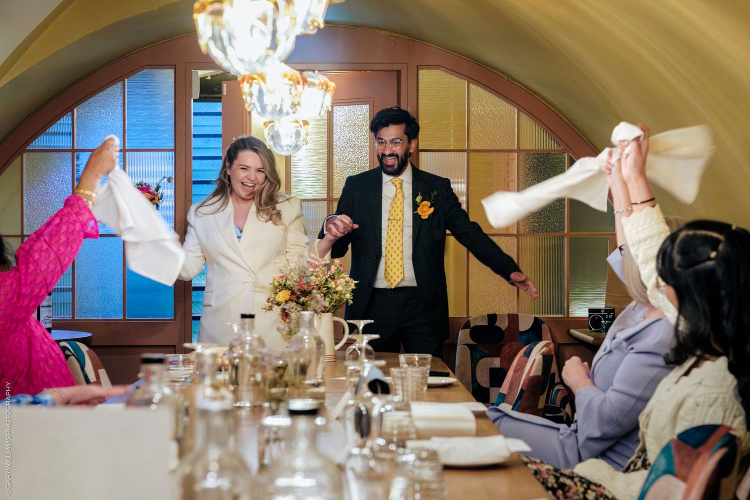 Bride and groom wave white napkins at wedding reception dinner table with guests in elegant venue.