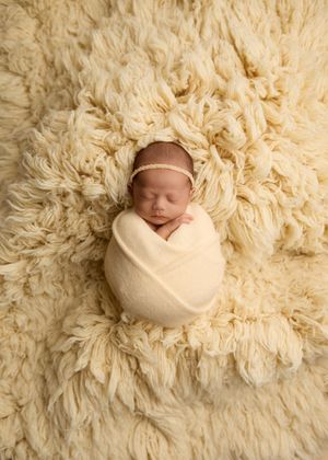 Newborn baby swaddled in cream wrap with gold headband, sleeping peacefully on fluffy ivory flokati rug. -- Sara Prue Photography Denver