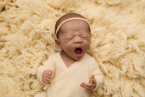 Newborn baby girl yawning on fluffy cream blanket wearing white wrap and gold headband. -- Sara Prue Photography Denver