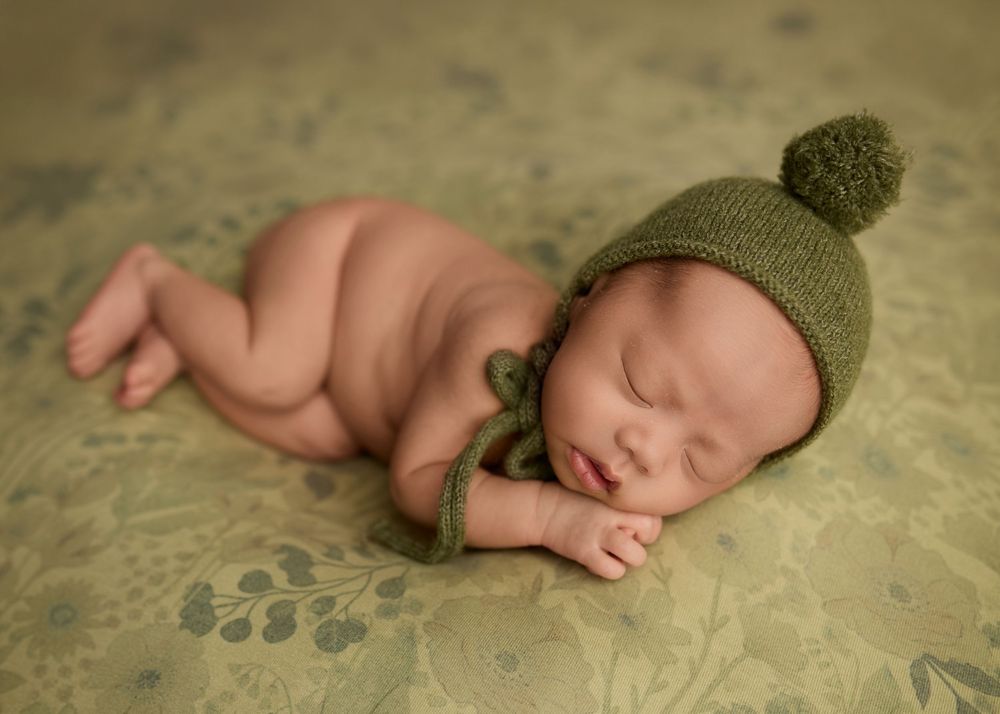 Sleeping newborn baby wearing a green knit pom-pom bonnet hat, posed on a floral green backdrop. -- Sara Prue Photography Denver