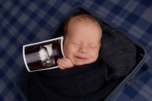 Smiling newborn baby wrapped in navy swaddle holding ultrasound photo, lying in basket on plaid blanket. -- Sara Prue Photography Denver